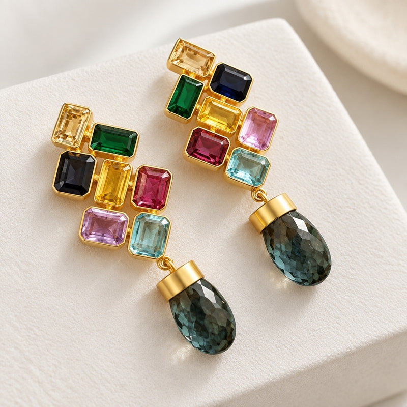 Gold-tone geometric earrings with multicolor rectangular accents and dark faceted teardrop dangles on an ivory display