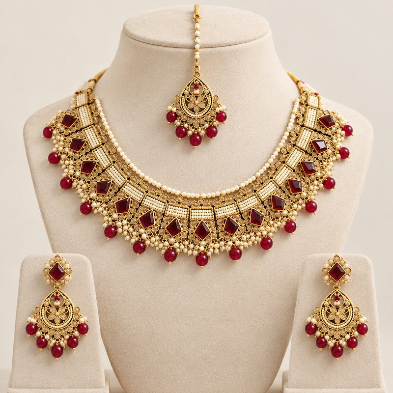 Red stone and gold-tone necklace set with pearl-style beadwork, matching drop earrings and maang tikka on a cream jewelry display.