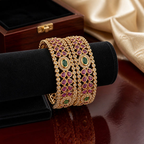 Gold-plated bridal bangles with ruby-colored stones, emerald green center accents, and American diamond detailing displayed on a black jewelry stand