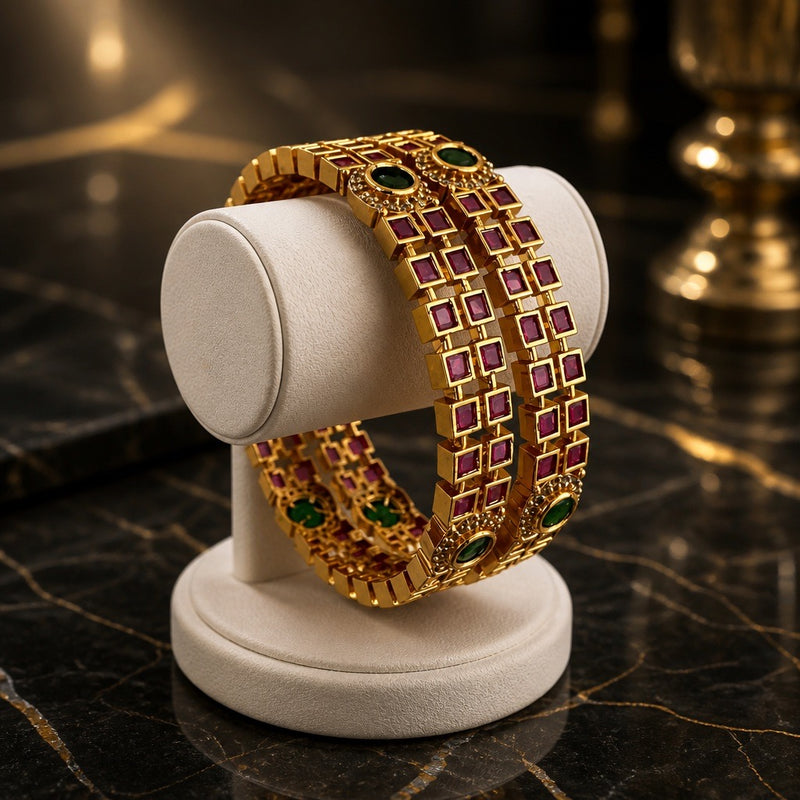 Gold-plated size 2.6 bangles with ruby red square stones and emerald green oval accents displayed on a cream jewelry stand
