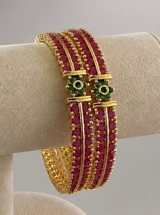 Gold-plated openable bangles with ruby red stones and green floral accents in size 2.8 displayed on a cream jewelry stand